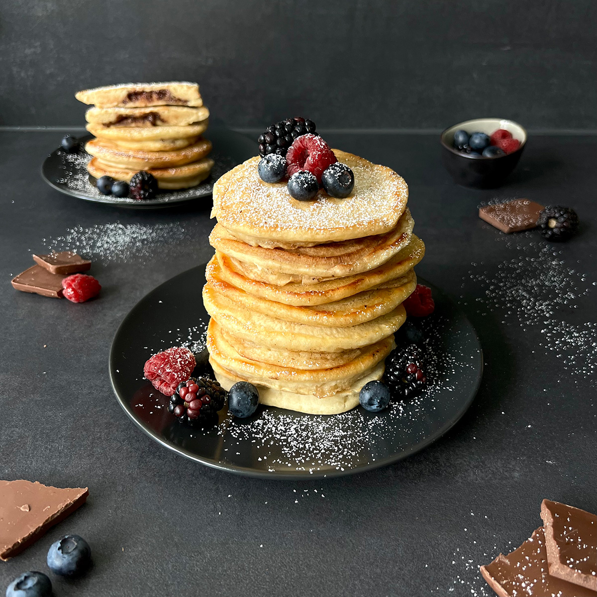 Das Bild zeigt Pancakes aufgestapelt auf einem dunklen Teller. Im Hintergrund sieht man ein weiteren Teller mit angeschnittenen Pancakes.