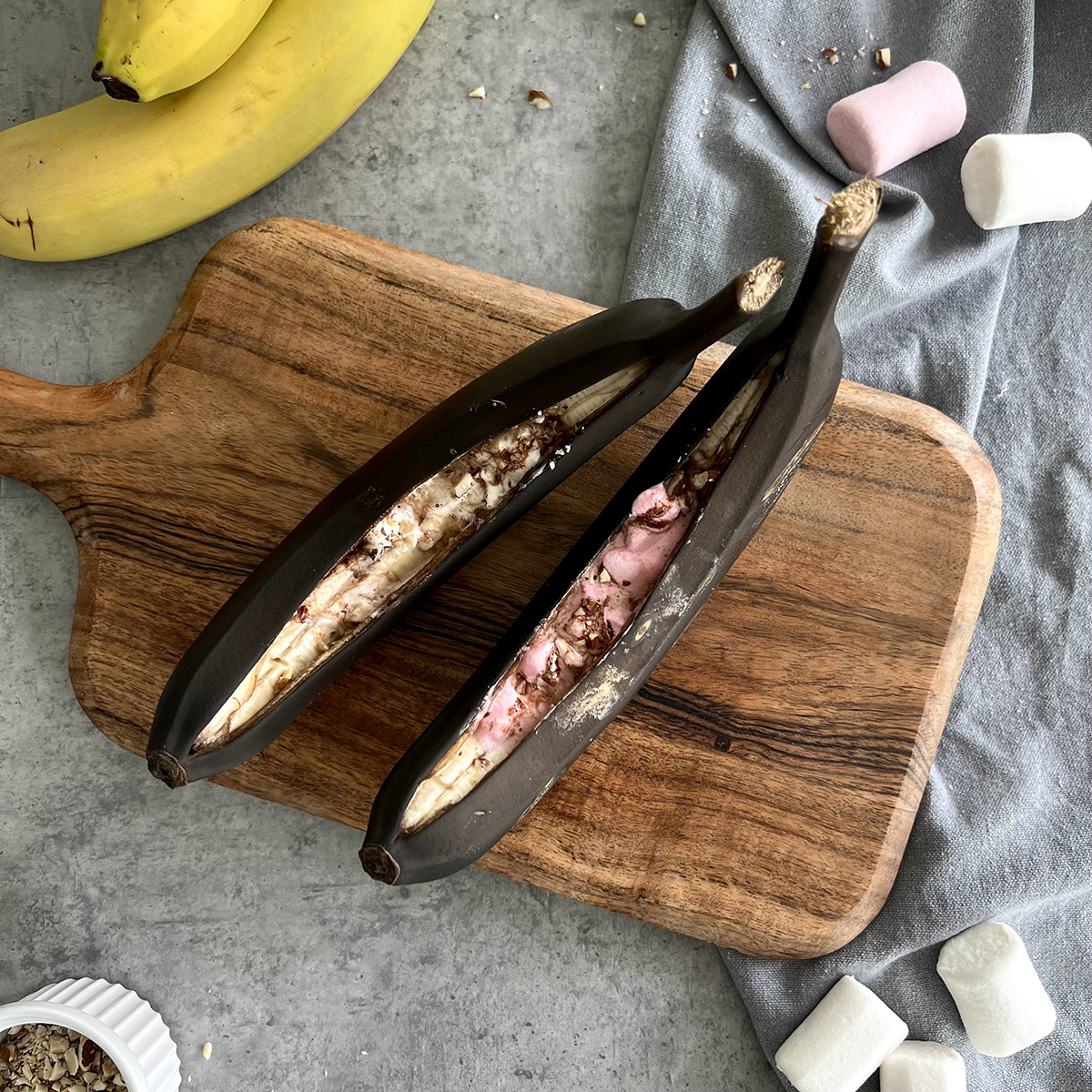 Grill Banane mit Marshmallows Zwei gegrillte Banane gefüllt mit Marshmallows auf einem Holzbrett. Die Bananen sind mit Schale und komplett schwarz vom Grillen.