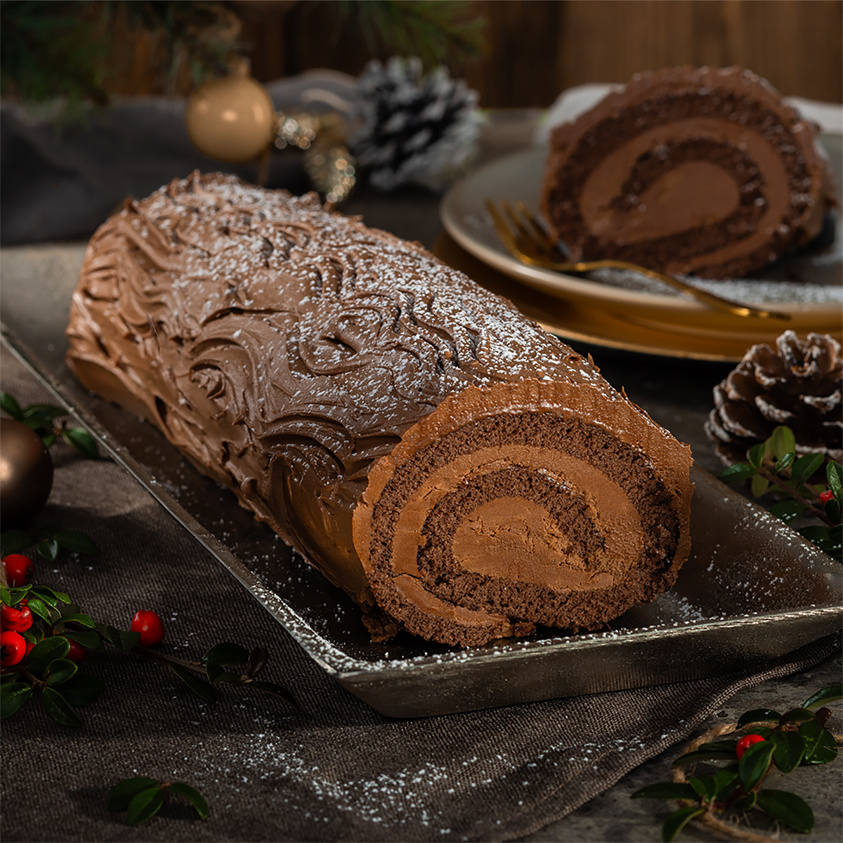 Das Bild zeigt eine dunkle Rolle Bûche de Noël auf einer länglichen dunklen Platte. Insgesamt ist es sehr dunkel dekoriert mit Tannenzapfen und Sträuchern.