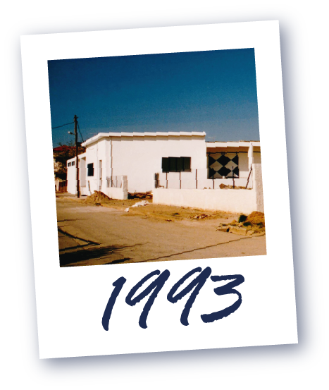 Weißes Gebäude an einer Straße unter klarem Himmel, darunter die Jahreszahl '1993'.