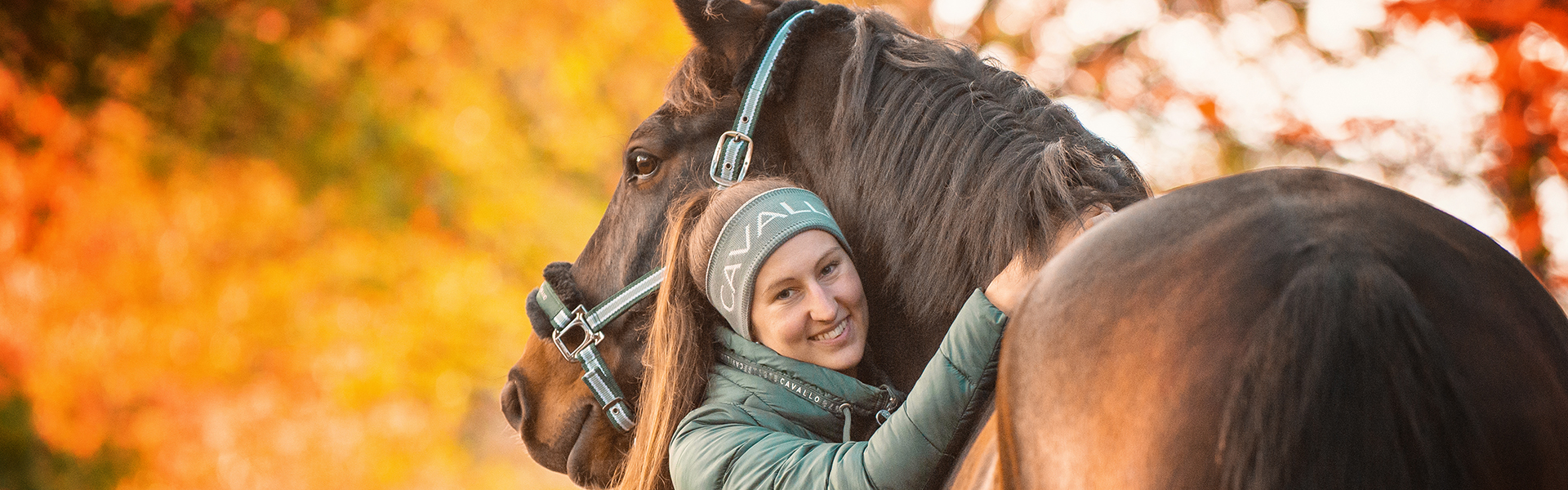 Lächelnde Frau umarmt ein braunes Pferd vor herbstlichem Hintergrund mit orangefarbenen Blättern.