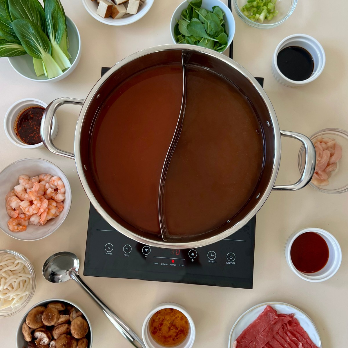 Auf einer Kochplatte steht ein zweigeteilter Hot Pot Topf gefüllt mit zwei verschiedenen Brühen. Drumherum sind verschiedene Zutaten verteilt.