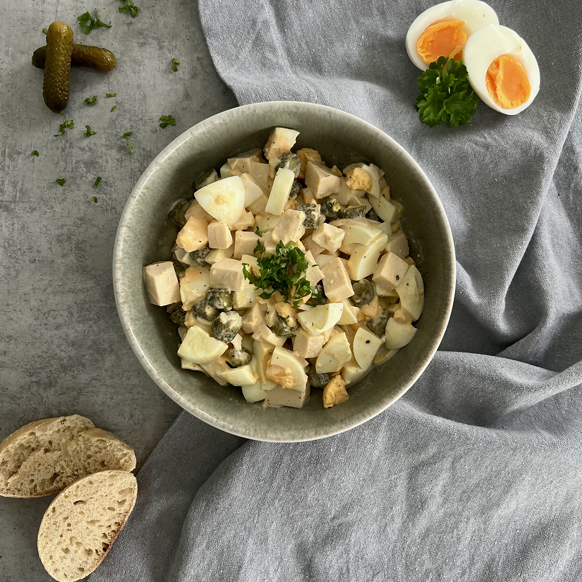Das Bild zeigt eine tiefe Schale mit Salat aus Eiern, Eierstich und Gewürzgurken. Serviert wird er mit Baguette.