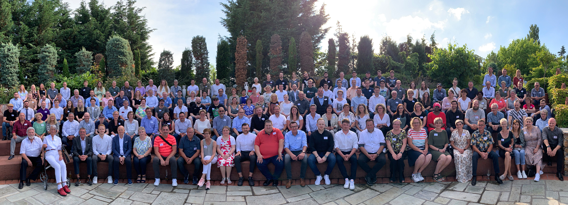 Großes Gruppenfoto, die auf mehreren Reihen im Freien sitzen und stehen, umgeben von Bäumen und Sträuchern.