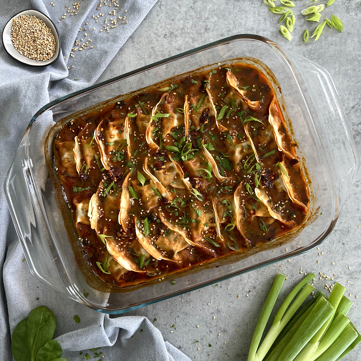 Das Bild zeigt eine durchsichtige Auflaufform gefüllt mit Dumplings. Sesamsamen und Lauch liegen daneben. 