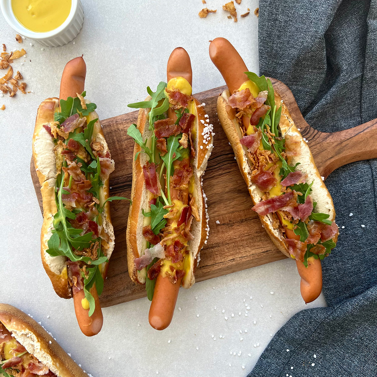 Das Bild zeigt 3 Hotdogs auf einem Brettchen. Ein weiterer wird angeschnitten und eine Schale Dip ist zu sehen.