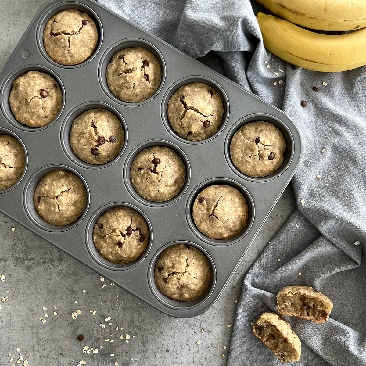 Das Bild zeigt zwölf Bananen Muffins in einer grauen Muffinform. Dazu zwei Bananen.