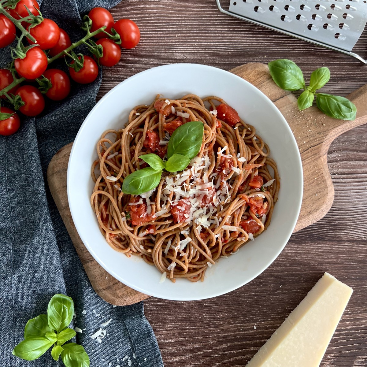 Die Spaghetti in Basilikum-Tomatensauce ist einem weißen Teller angerichtet, dieser steht auf einem Holzbrett, das Gericht ist mit Basilikum Blätter verfeinert