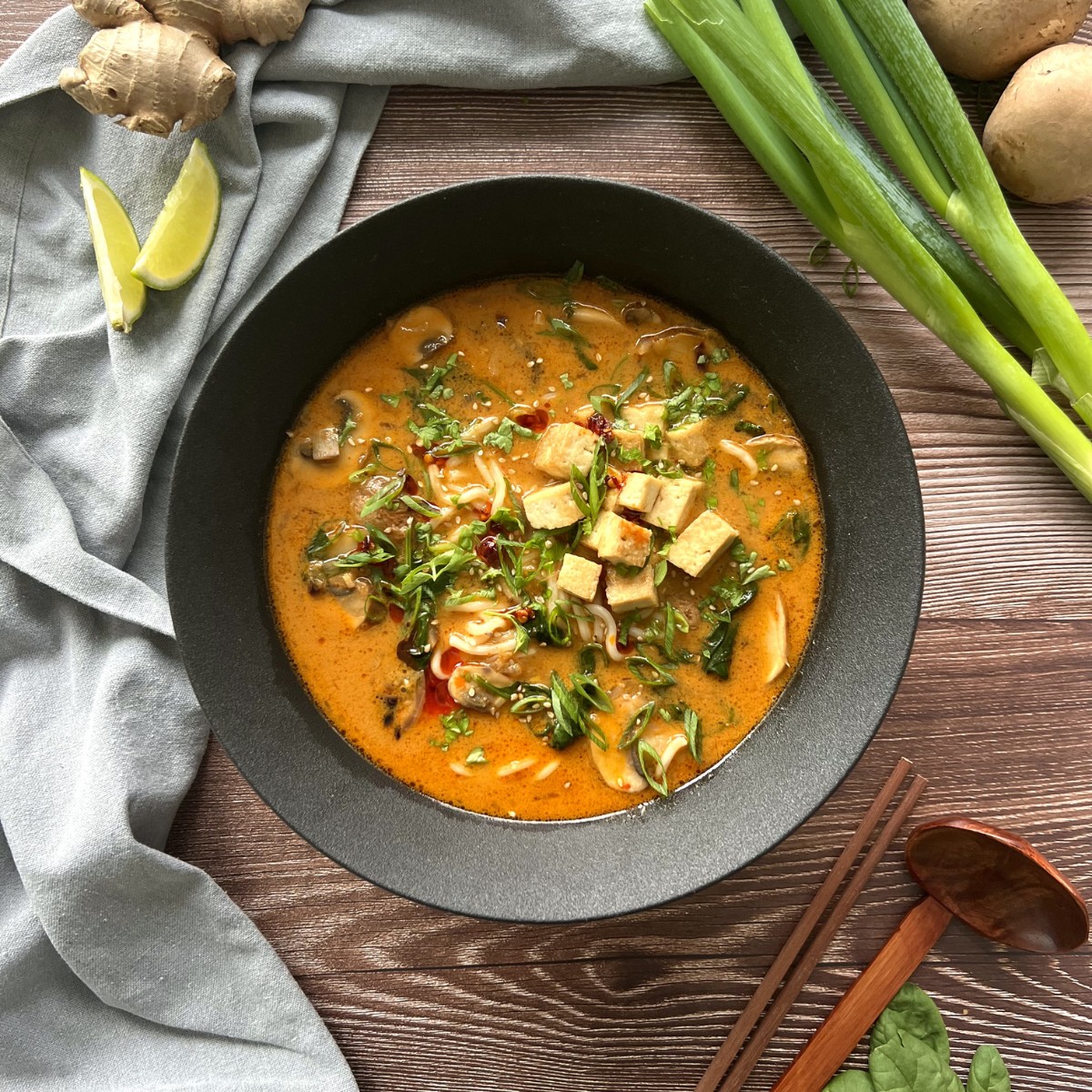 Das Bild zeigt in der Mitte des Bildes die Ramen Suppe in einer schwarzen tiefen Schale. Daneben sind Limettenspalten, Frühlingszwiebeln, Ingwer und Essstäbchen.