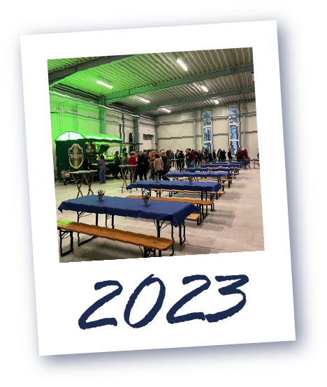 Große Halle mit langen Tischen und Bänken, Menschen stehen im Hintergrund, darunter die Jahreszahl '2023'.