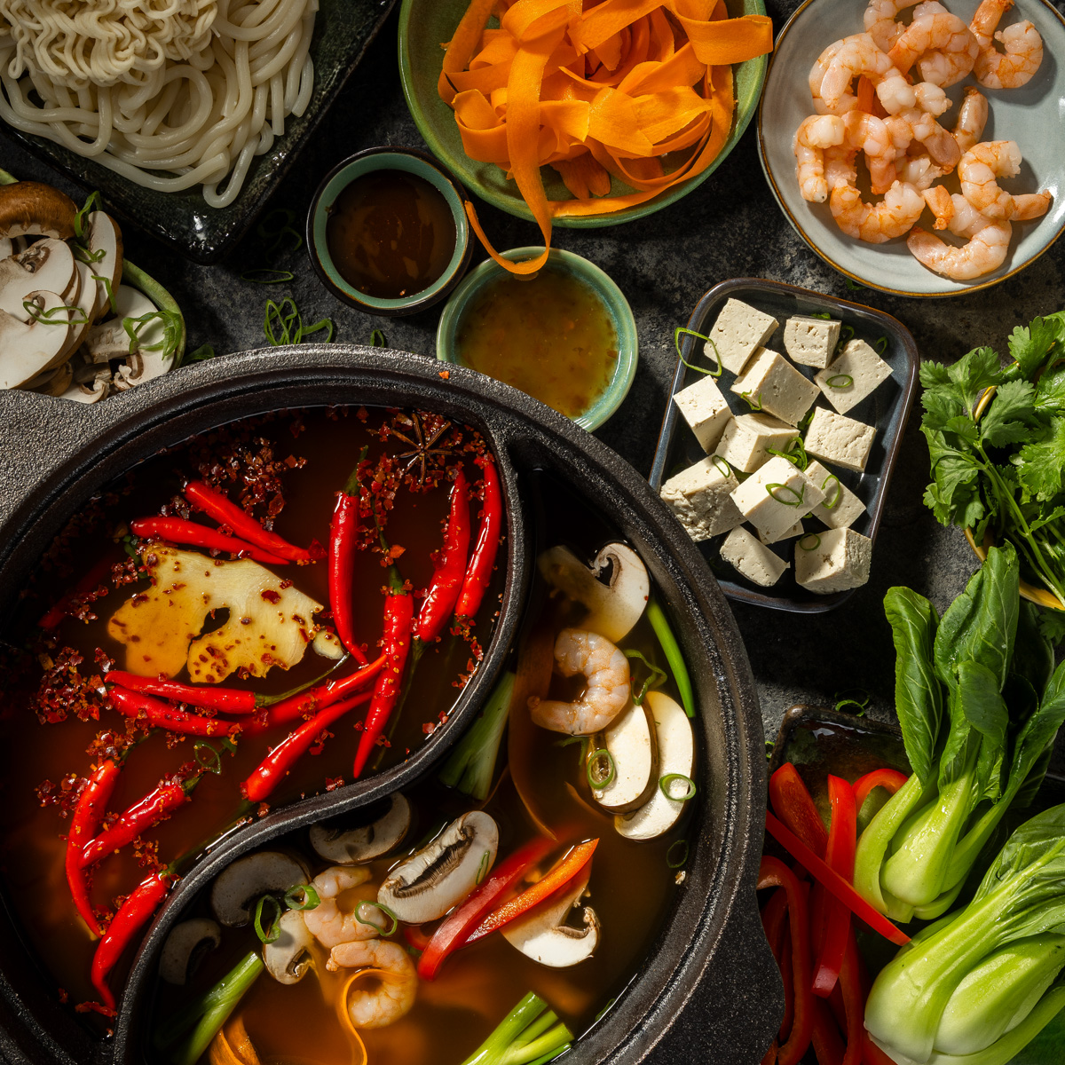 Die Hot Pot Brühen sind in zwei Fächer aufgeteilt und mit Zutaten befüllt. Unter anderem Chili und Pilze. Drum herum sind Hot Pot Zutaten in Schalen verteilt.