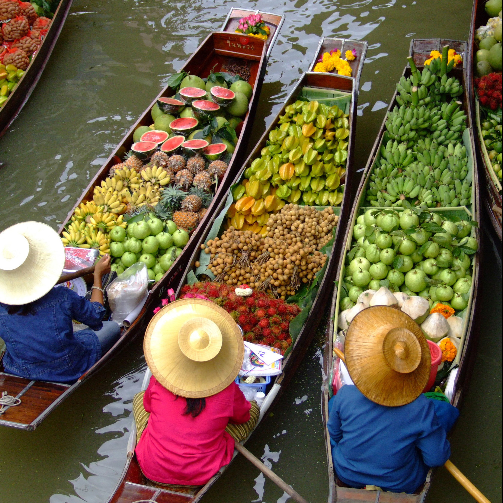 Drei Boote denen verschiedene Früchte liegen. Drei Personen mit Strohhüten steuern die Boote.
