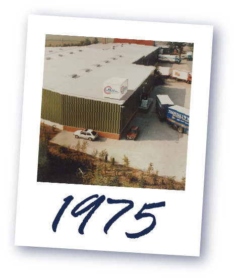 Luftaufnahme eines großen Industriegebäudes mit mehreren Lastwagen auf dem Gelände, darunter die Jahreszahl 1975.