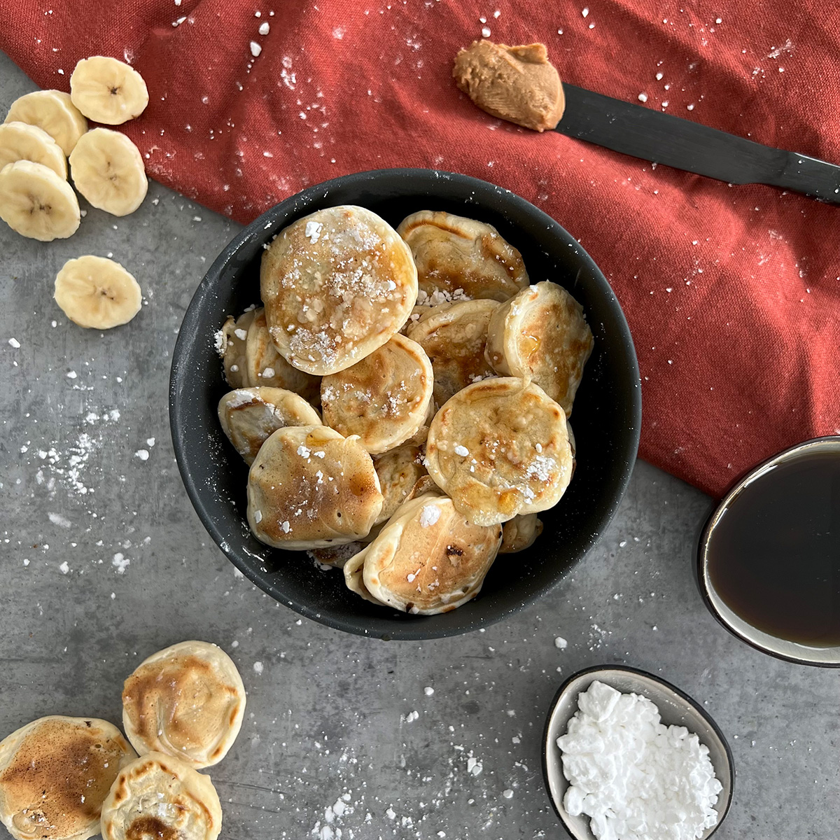 Das Bild zeigt mehrere kleine Pancakes in einer tiefen Schale. Sie sind mit etwas Puderzucker überzogen und eine Schale mit Schokosauce steht daneben.