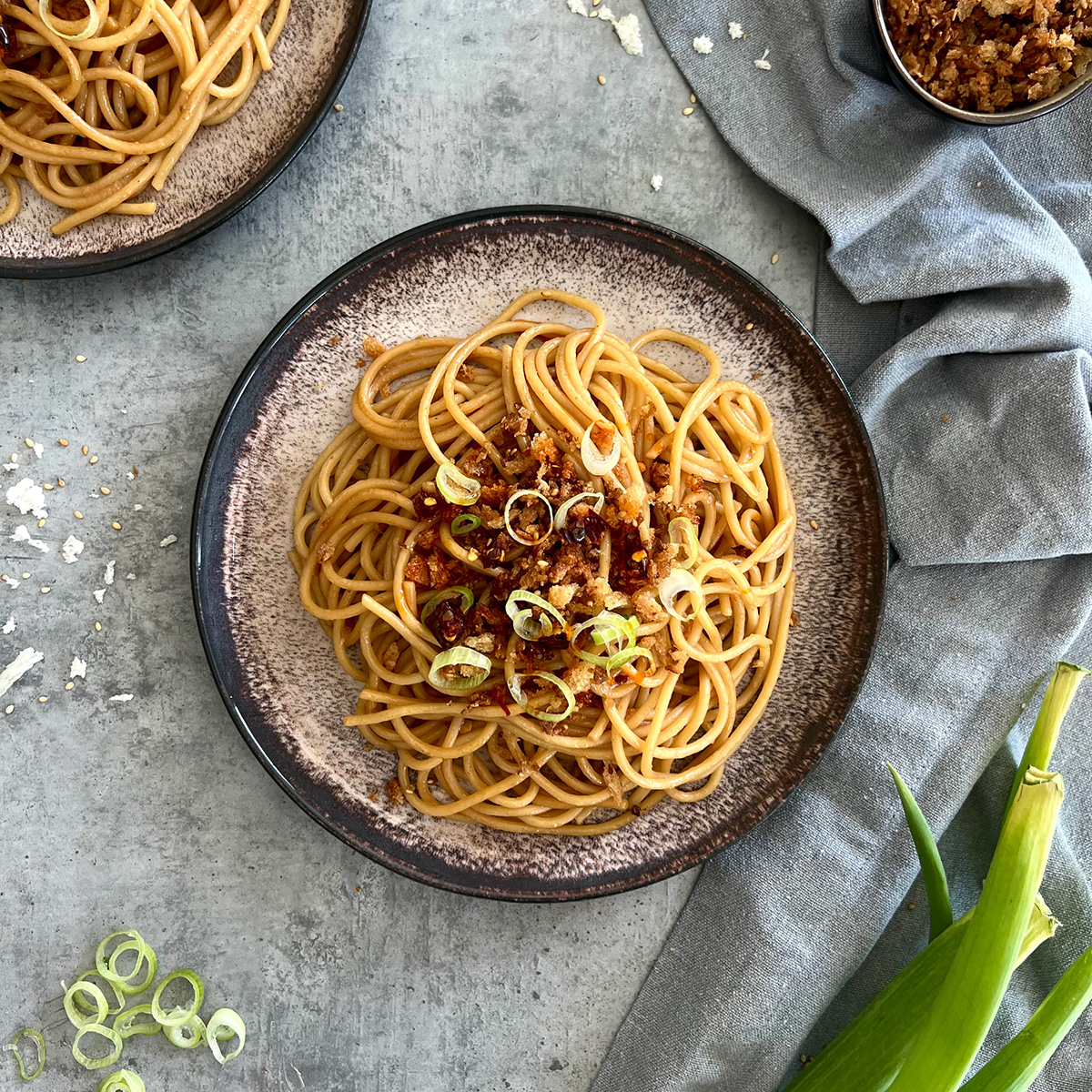 Das Bild zeigt einen Teller gefüllt mit Spaghettis in der Butter. Getoppt mit Crunch und serviert mit Lauchzwiebel. Ein zweiter Teller ist angeschnitten.