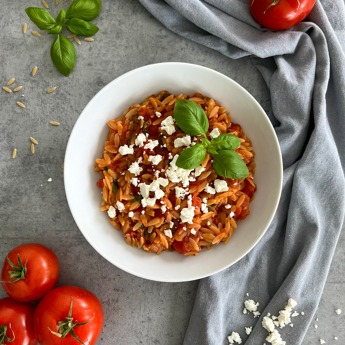 Das Bild zeigt eine weiße Schale gefüllt mit den Kritharaki. Es werden Tomaten, Feta und Basilikum angeschnitten.