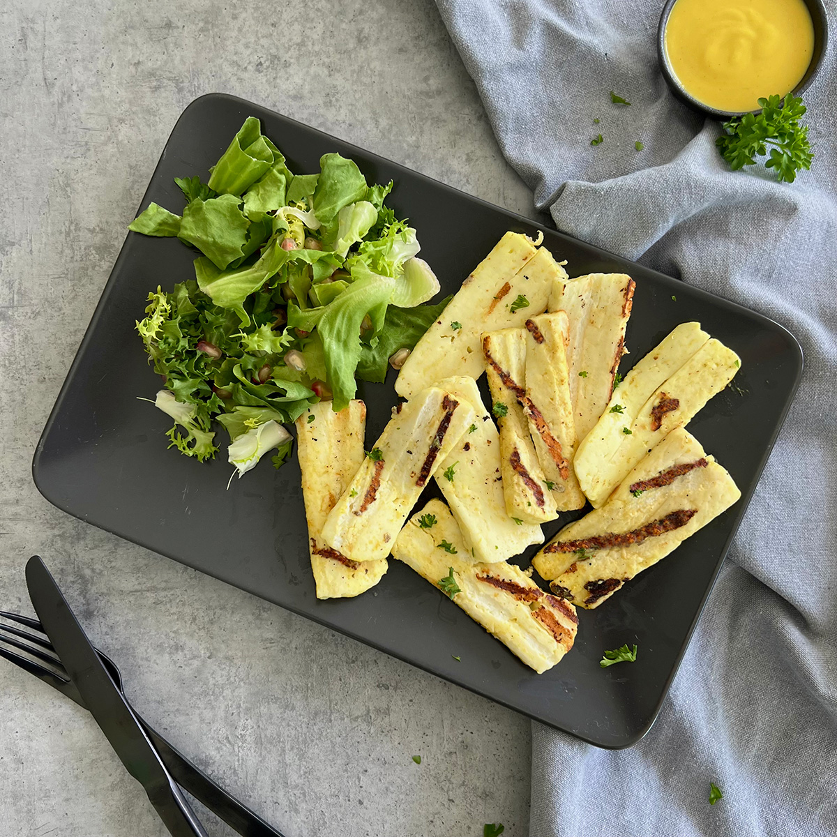 Gegrillter Halloumi in Honig Senf Sauce Gegrillte Halloumi Scheiben liegen neben etwas grünem Salat auf einem schwarzen rechteckigen Teller. In einer kleinen Schale ist etwas Sauce.