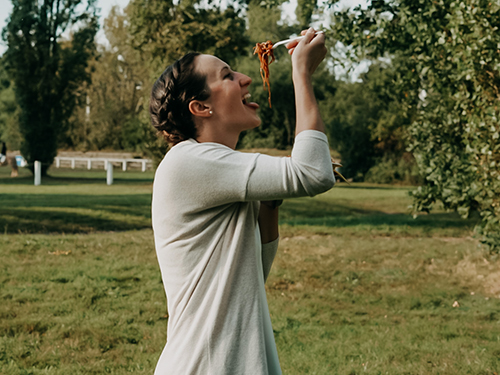 Eine Frau in der Mitte, die im grünen Park mit Stäbchen Spaghetti isst. isst, umgeben von Bäumen und Wiese.