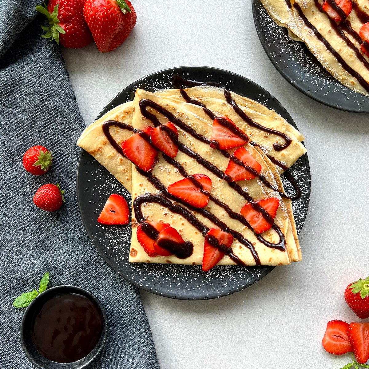 Zwei gefaltete Crêpes auf einem Teller getoppt mit Schokoladensauce, Erdbeeren und Puderzucker.