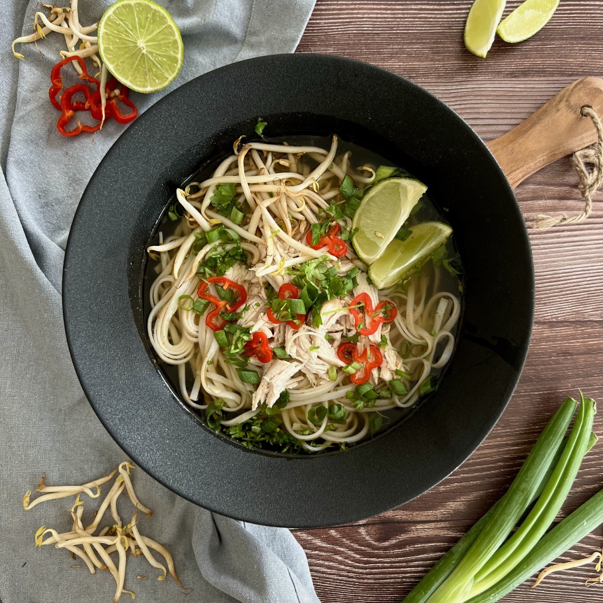 Das Bild zeigt in der Mitte des Bildes eine Pho Ga. Zur Deko befinden sich neben der Schüssel Limetten, Frühlingszwiebeln, Chili Scheiben und Sprossen.